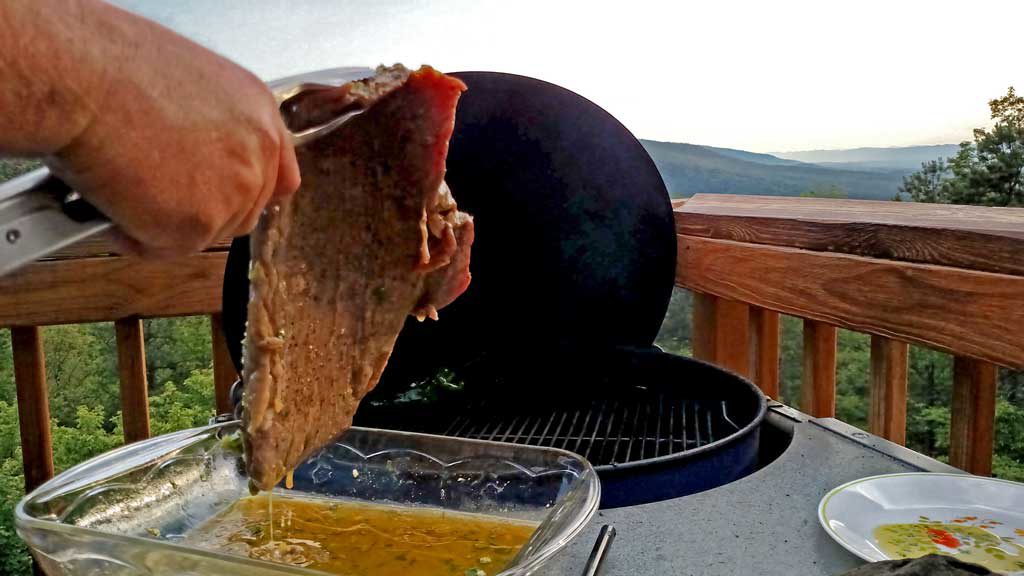 hand holding flank steak with tongs beside grill with mountain view