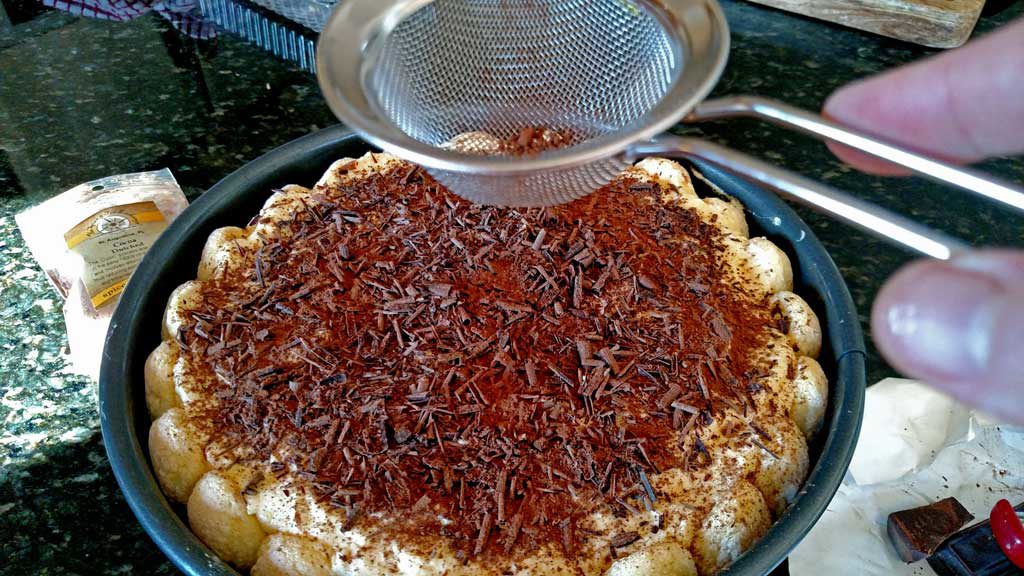 dusting top of homemade tiramisu with coco powder and chocolate shavings