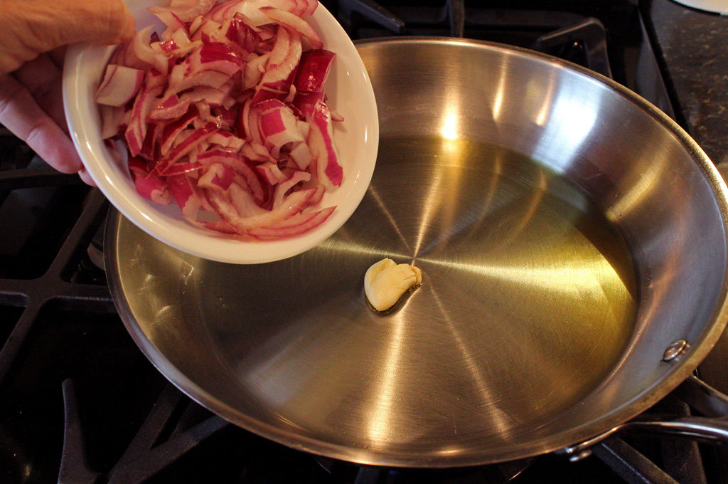 garlic and oil in skillet with bowl of onions getting ready to be tossed in