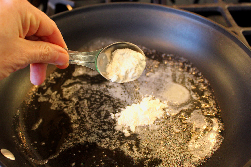 measuring spoon of flour added to skillet