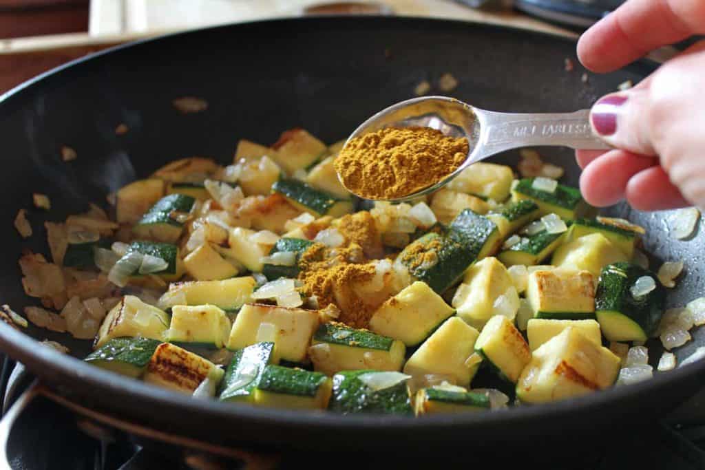 Curry in a measuring spoon being added to the pan of zucchini and onions.