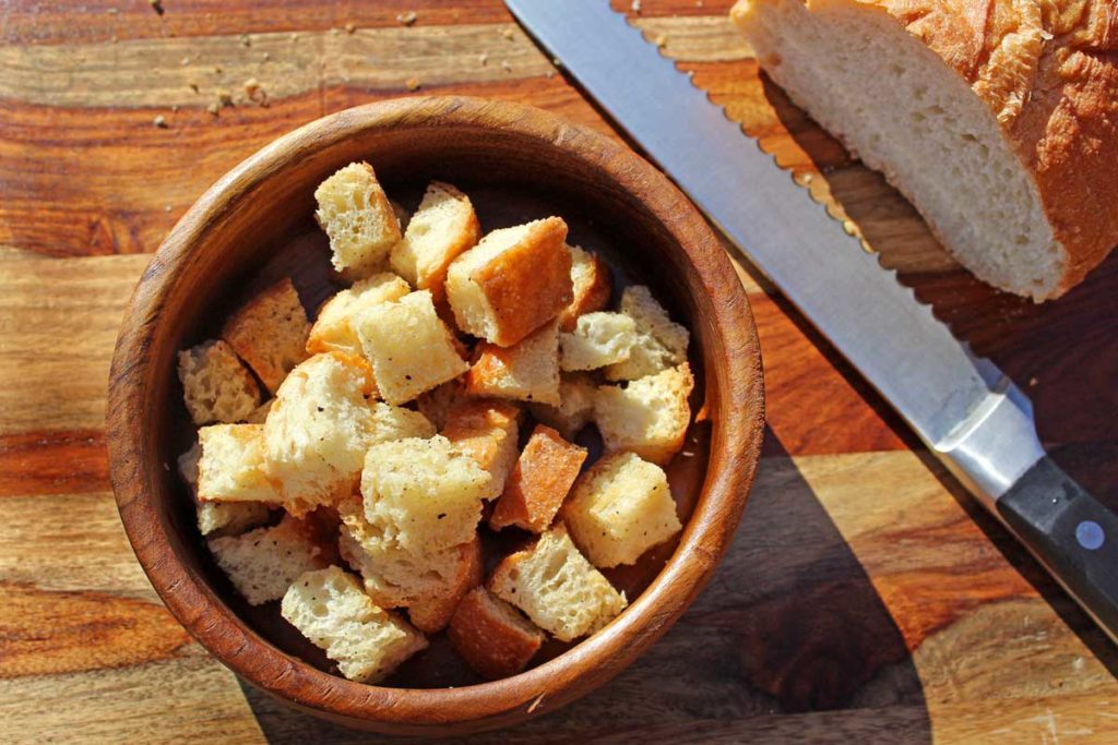 A bowl of croutons beside a bread knife and a loaf of bread.