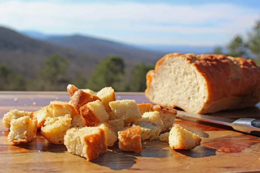 A loaf of bread with homemade croutons on a board with the mountains in the background.