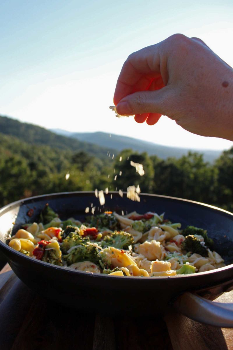 sprinkling parmesan cheese into the pan with the mountains in the background