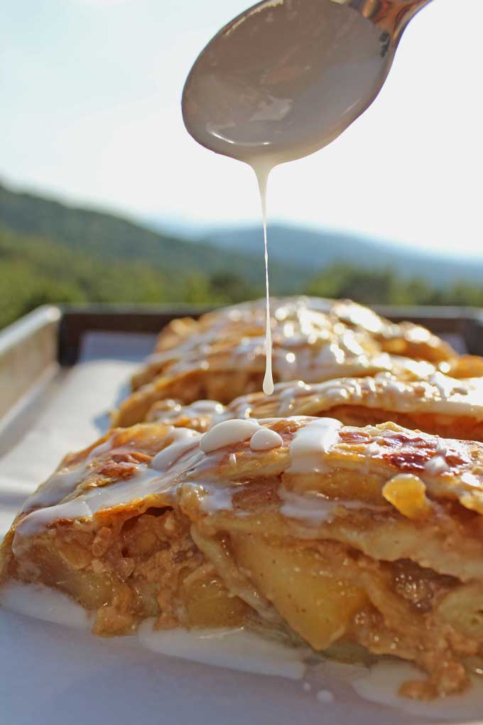 drizzling vanilla glaze onto the caramel apple strudel with mountain view in background
