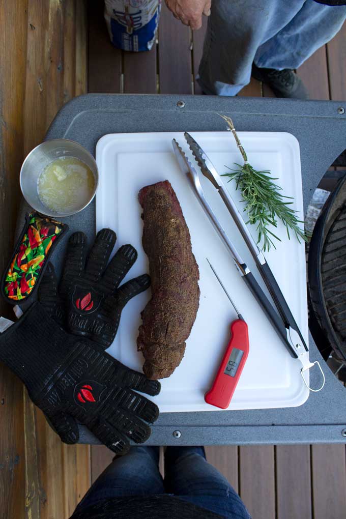 smoked meat on cutter board with tongs, herb brush, grill gloves, instant-read thermometer and pot of garlic butter
